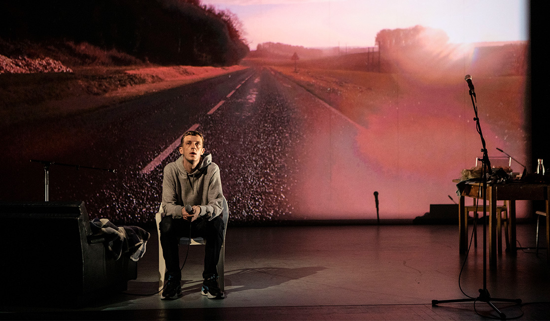 A man wearing a grey jumper and black pants sits on stage in front of a pink-toned backdrop of a road.