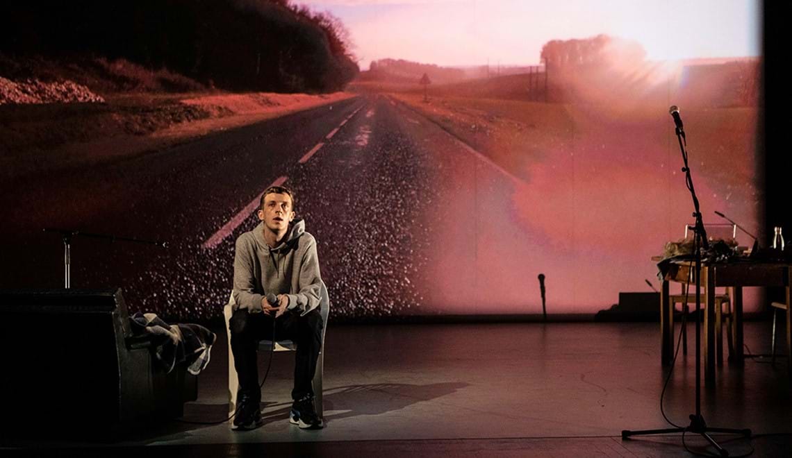 A man wearing a grey jumper and black pants sits on stage in front of a pink-toned backdrop of a road.