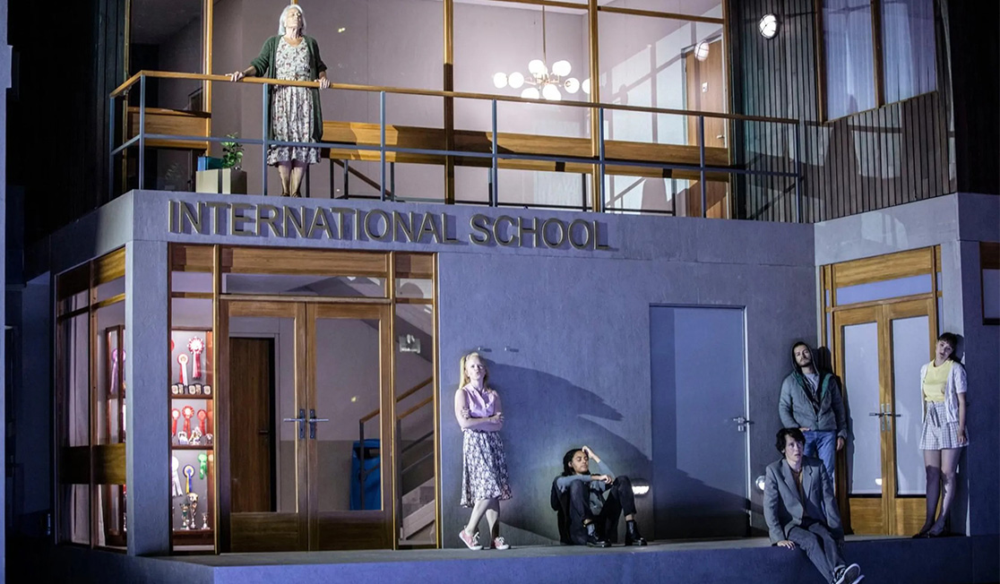 A woman with grey hair is wearing a dress and stands on a balcony. Below there are five students sitting and standing casually in front of a grey wall, they look tired. Letters above the door say 'INTERNATIONAL SCHOOL'