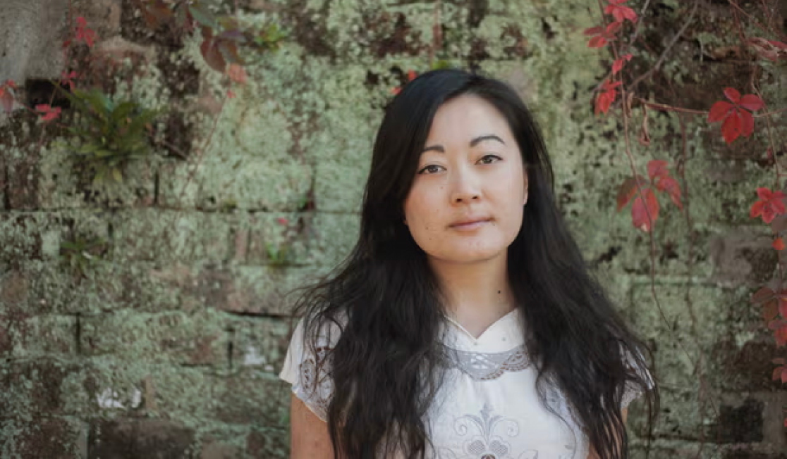 Jessie Tu stands in front of a dark brick wall. Jessie wears a white shirt and has long dark hair. Jessie is smiling slightly.
