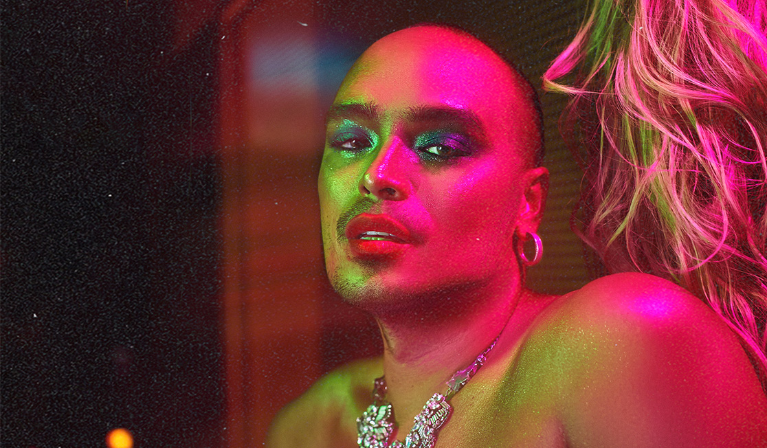 Hedwig is standing in front of a wall with a blonde wig hung up on it. They are shown in a vibrant pink light and wear a large silver necklace and a denim top with bare shoulders. They have hoop earrings on and seem to be sparkling.