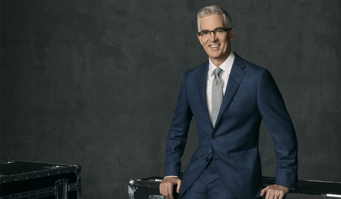 David Speers has grey hair and glasses and wears a white shirt, navy suit and silver tie, on a dark grey background 