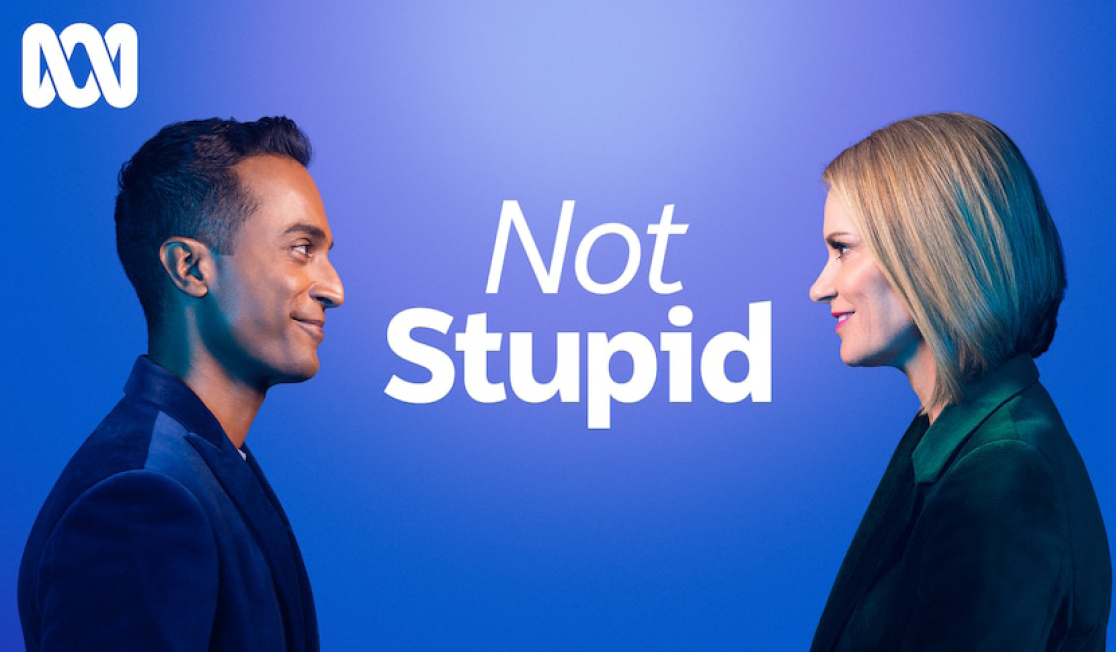 Two people stand facing each other with the words - Not Stupid, in the space between them. There is a blue background and both people wear dark clothes. One has a blonde bob and the other short dark hair. They are smiling.