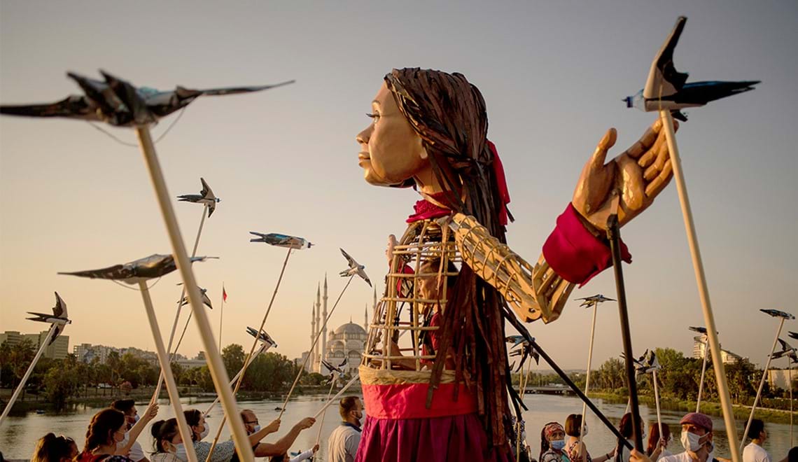 A 3.5 metre puppet of a young girl with brown hair and a red and purple skirt stands with her hands out. The puppet's chest is a cage and a puppet operator can be seen through the slats of the cage. People surround the puppet, quite a lot shorter than her, and look up at her. Many of the people are holding paper birds on long sticks, which rise up into the sky beside her.