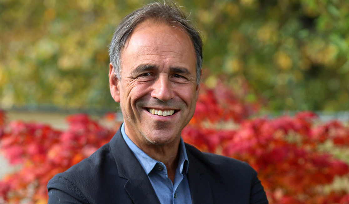 Anthony Horowitz wears a blue collared shirt and navy blazer, with red flowers pictured behind him 