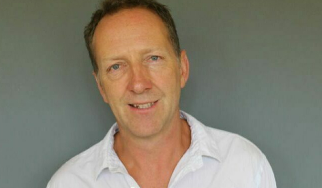 Tom Baldwin wears a white shirt and stands in front of a grey backdrop. He has short dark hair and is smiling.