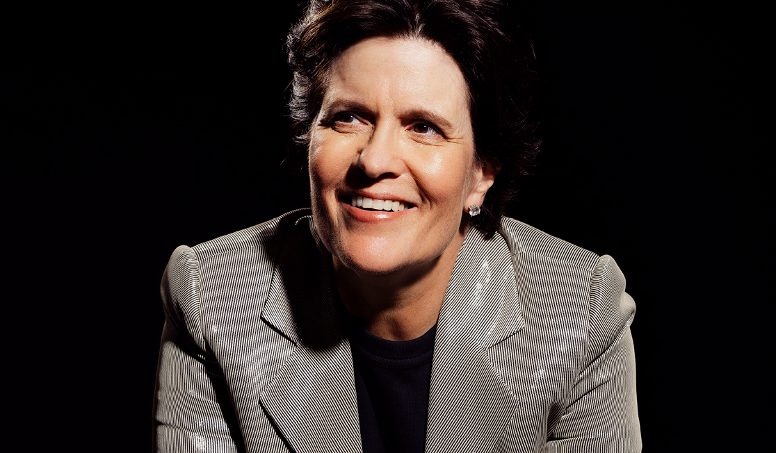 Kara Swisher wears a metallic grey suit jacket with a black top. Kara has short dark hair and is smiling in front of a black background.