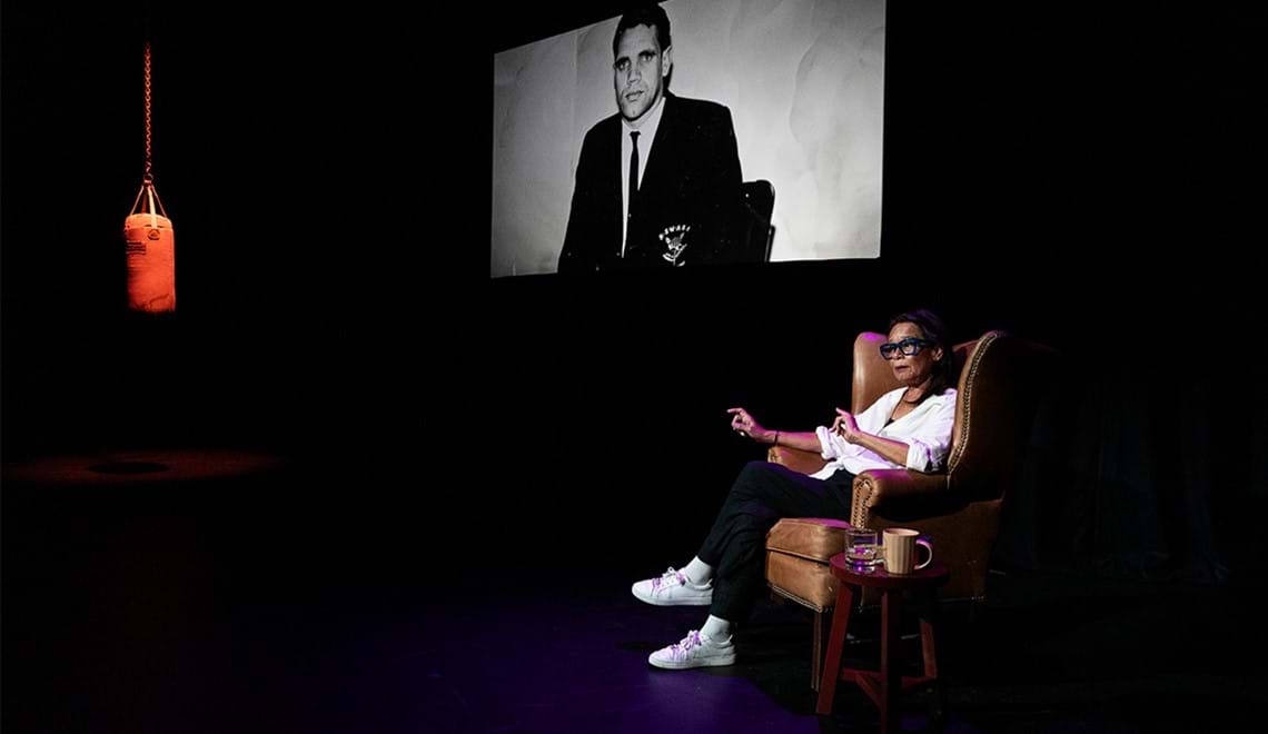 Rhoda Roberts sits in a brown leather armchair wearing a white shirt, black pants, white sneakers and glasses with black frames. on a screen behind the armchair there is a black and white image of a man in a suit. To the left of the stage there is a red punching bag hanging from the ceiling.