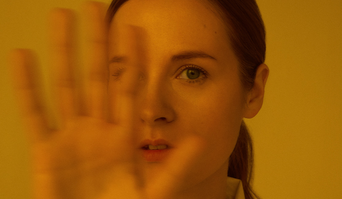 Hania Rani has a hand up in front of their face. They have brown hair tied in a ponytail. They are looking directly at the camera with a serious expression.