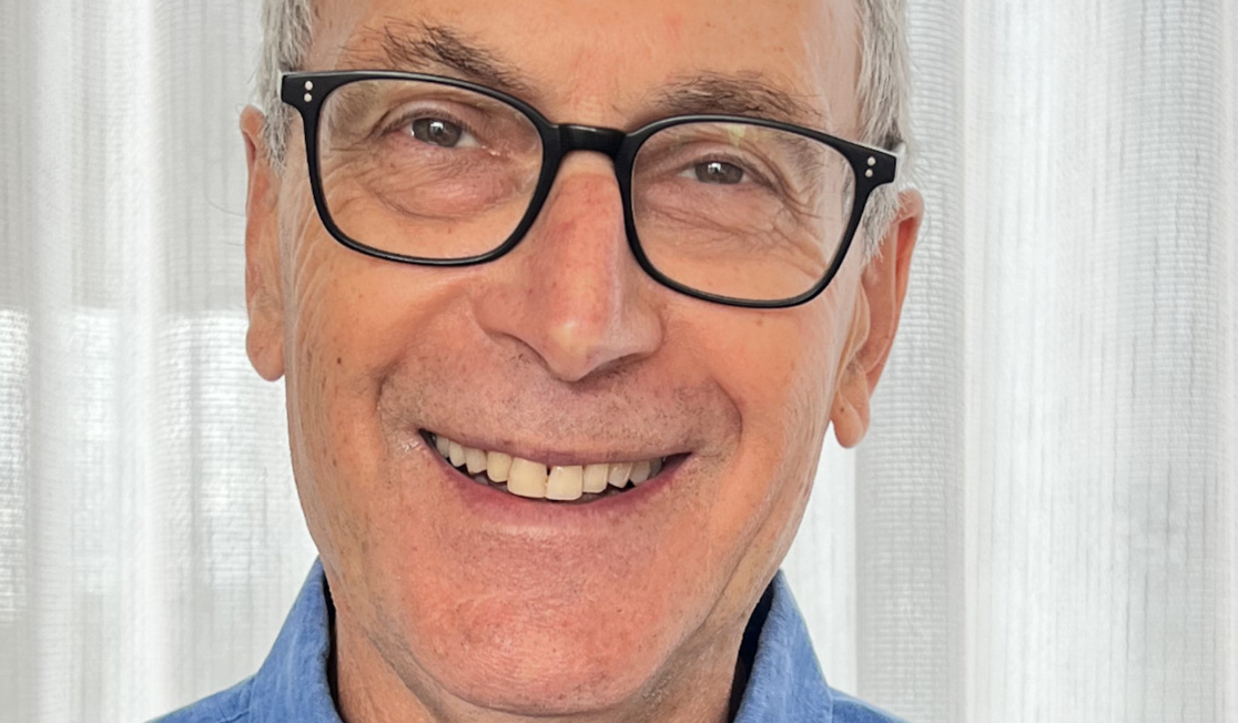 Eric Beecher wears a blue shirt and dark framed glasses. Eric is smiling in front of a white curtain.