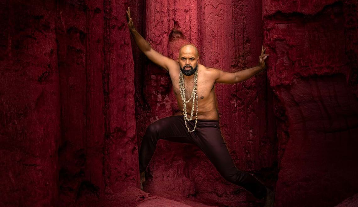 A shirtless man stands in front of a red cliff. He has his legs and arms spread wide apart and is looking at the camera with an intense, serious expression.