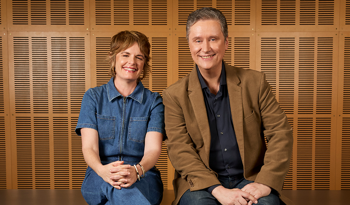 Two people sit on top of a black surface, the woman on the left is wearing a denim jumpsuit and gold bangles, she has short hair and is smiling. On the right, a man in a brown jacket and black shirt. He has short grey hair and is smiling.