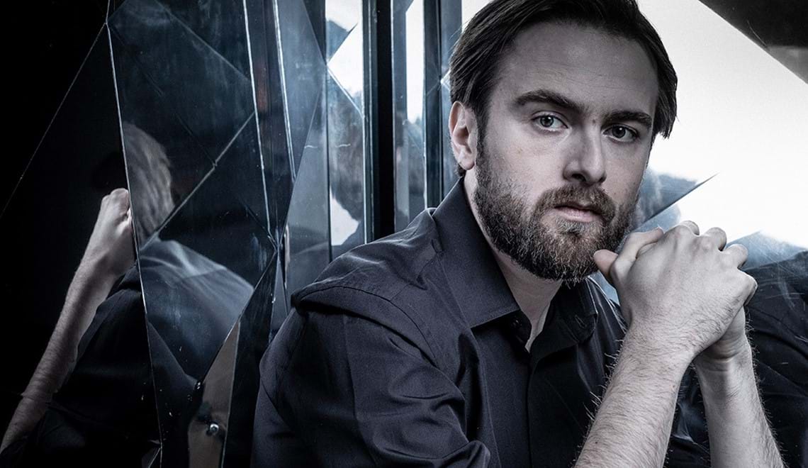 Daniil Trifinov clasps both hands in front of his chin and has a serious expression. He is surrounded by mirrors in geometric shapes. He wears a black shirt and has brown short hair and a beard.