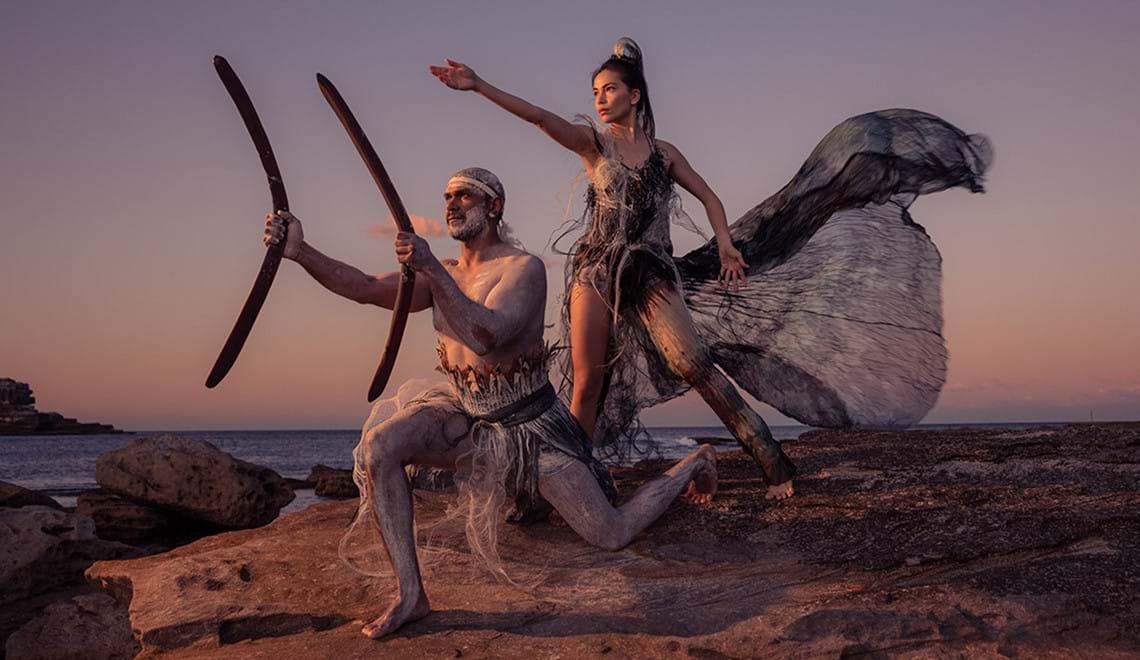 Two people stand on a rocky outcrop with the ocean visible behind them. They each strike a dramatic pose pointing towards something to the left of the camera.