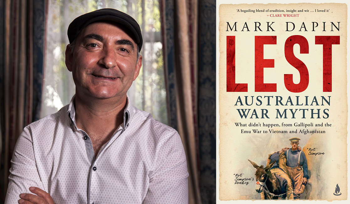 Mark Dapin wears a white shirt and a black hat. Mark stands in front of an open window and is smiling.