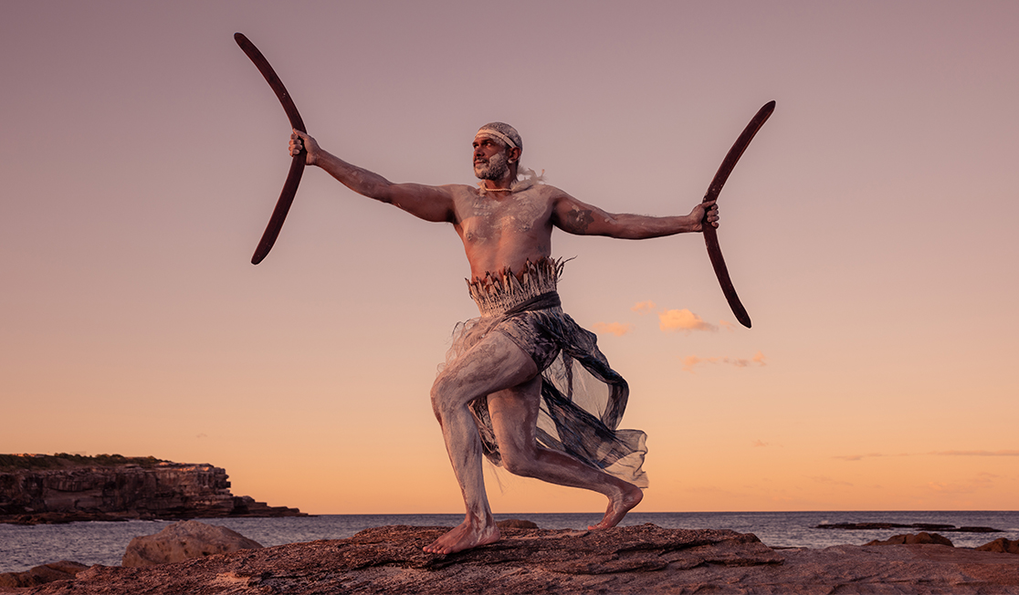 A man stands on a cliff with his arms reaching out wide. He is holding a boomerang in each hand. The ocean and a distant bluff are visible behind him..