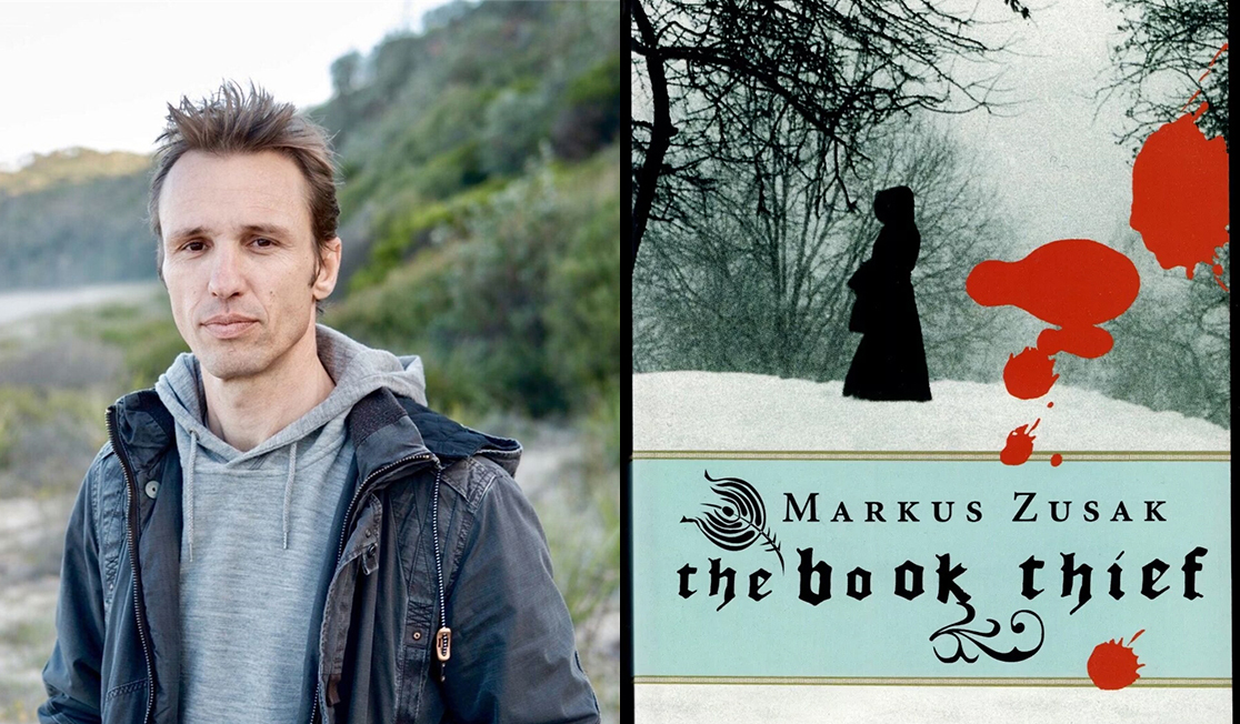 Markus Zusak wears a grey sweatshirt and a blue jacket. He is outdoors with green bushes pictured behind him.