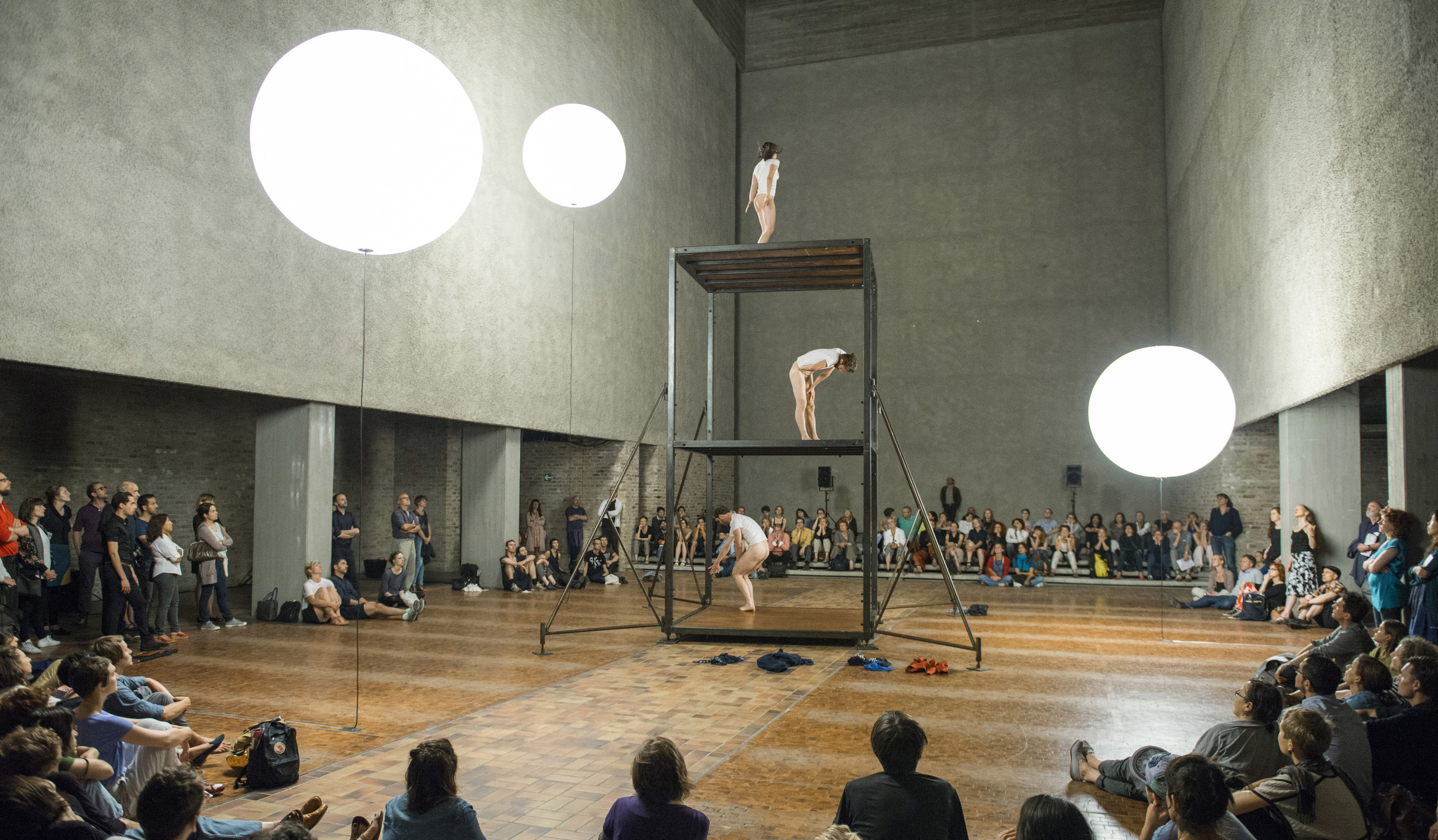Three dancers are standing on a structure, similar to scaffolding, it has 3 levels and the dancers are wearing white t-shirts and no pants. There is a seated audience surrounding them.