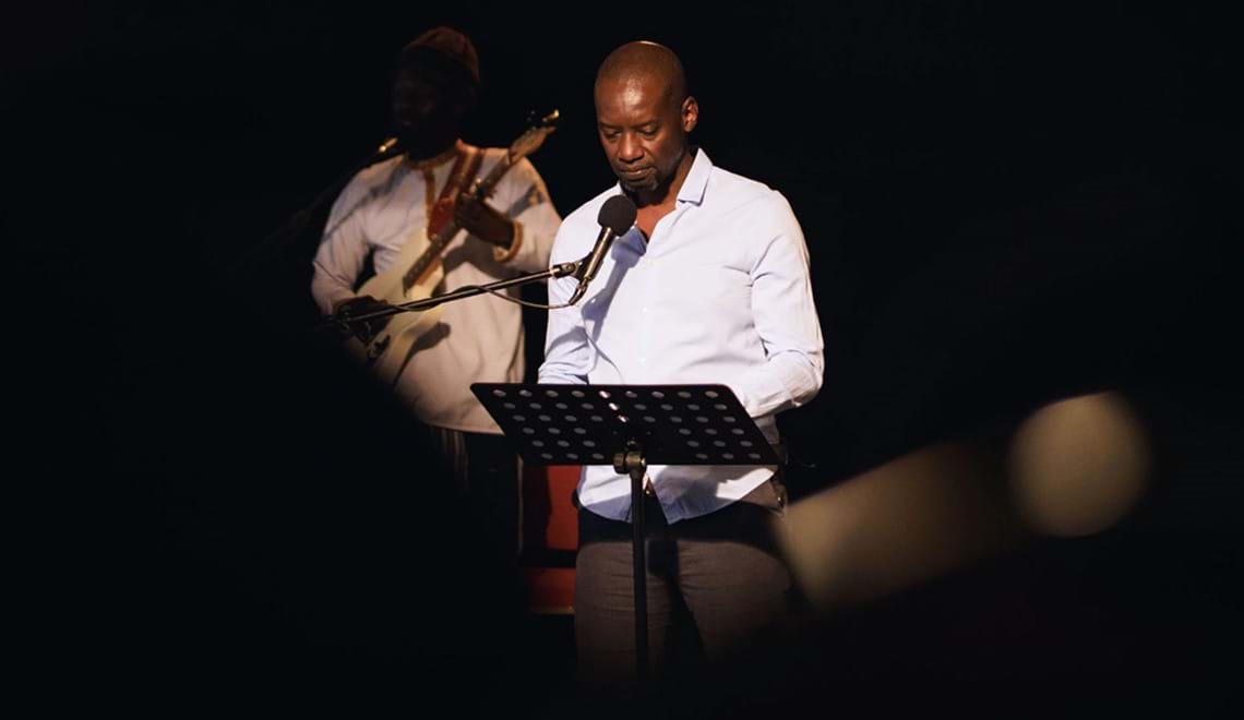 A man stands in front of a music stand, there is a microphone stand pointed towards him. He wears a white shirt and is surrounded by darkness. There is a guitarist