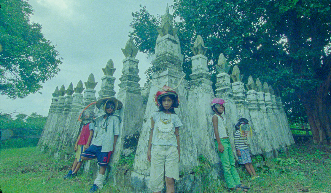 Five young people in colourful hats and clothing stand in front of several grey stone sculptures in a lush setting.