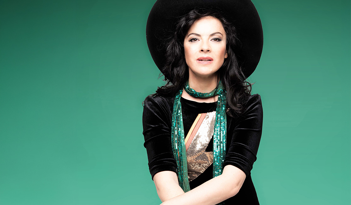 Camille O'Sullivan sits in front of a green background wearing a black top, a sequin green scarf and a wide brimmed black hat. Her arms are resting, crossed in front of her and she is looking straight ahead with her mouth open.