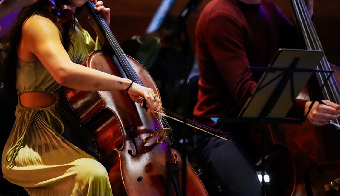A musician is playing the cello on stage. They are wearing a green dress and have long brown hair but we cannot see their face. Their arm is raised to play the cello with a bow. There is a music stand in front of them and another cello player in the background.