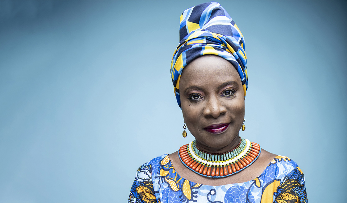 A woman wearing a blue and yellow dress and matching head wrap smiles at the camera in front of a plain blue background.