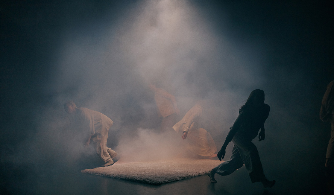 Four people dressed in beige clothing are walking away from a shaggy white rug on a stage. There is a light with smoke coming over the rug, but the people are half in darkness.