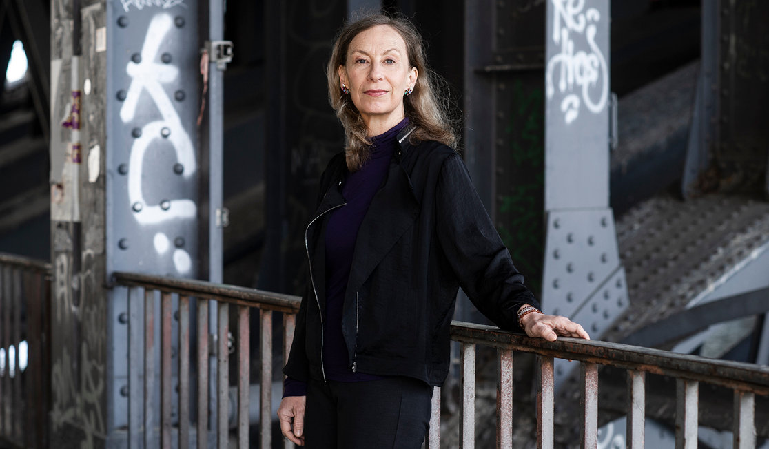 A woman stands with one hand on the railing of a bridge. She has long, light brown hair and wears a black jacket and pants and a navy top. She is smiling.