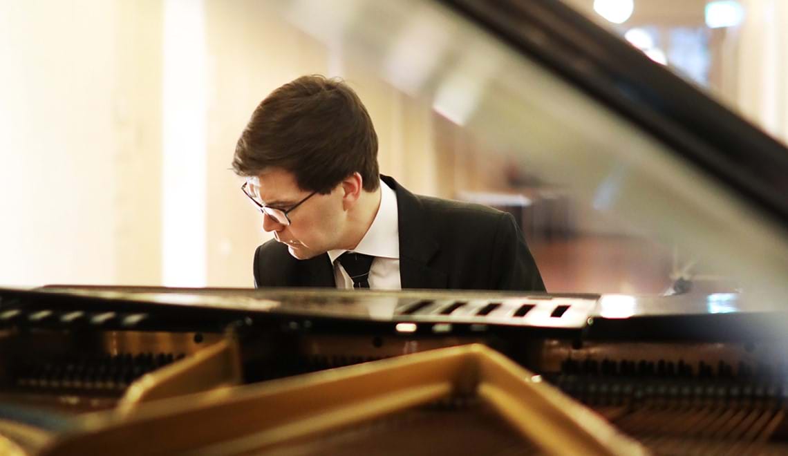A musician sits at a piano wearing a black suit and tie and a white shirt. We are looking at him through the open top of the piano. He wears glasses and has short brown hair.