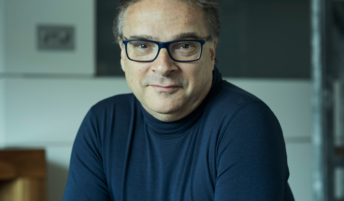 Orlando wears a dark blue, long-sleeved top, dark glasses and has short grey hair