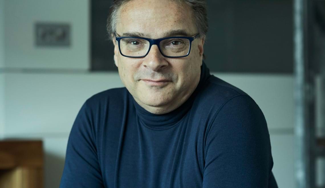 Orlando wears a dark blue, long-sleeved top, dark glasses and has short grey hair