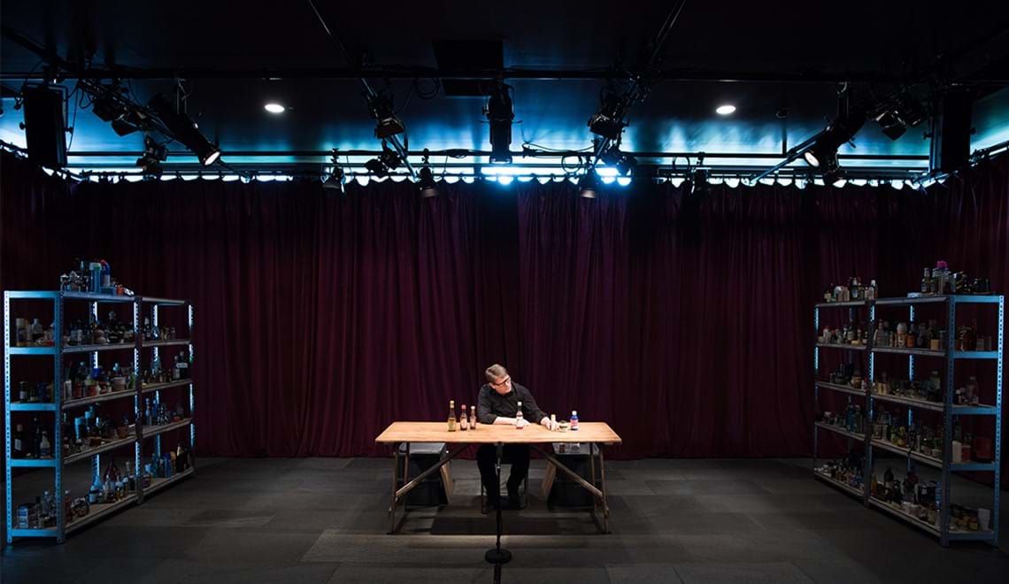 A person wearing black sits at a trestle table. They are spotlighted in a dark room. They have sauce bottles and various condiments in front of them, they have their hand reached out to hold one of them.