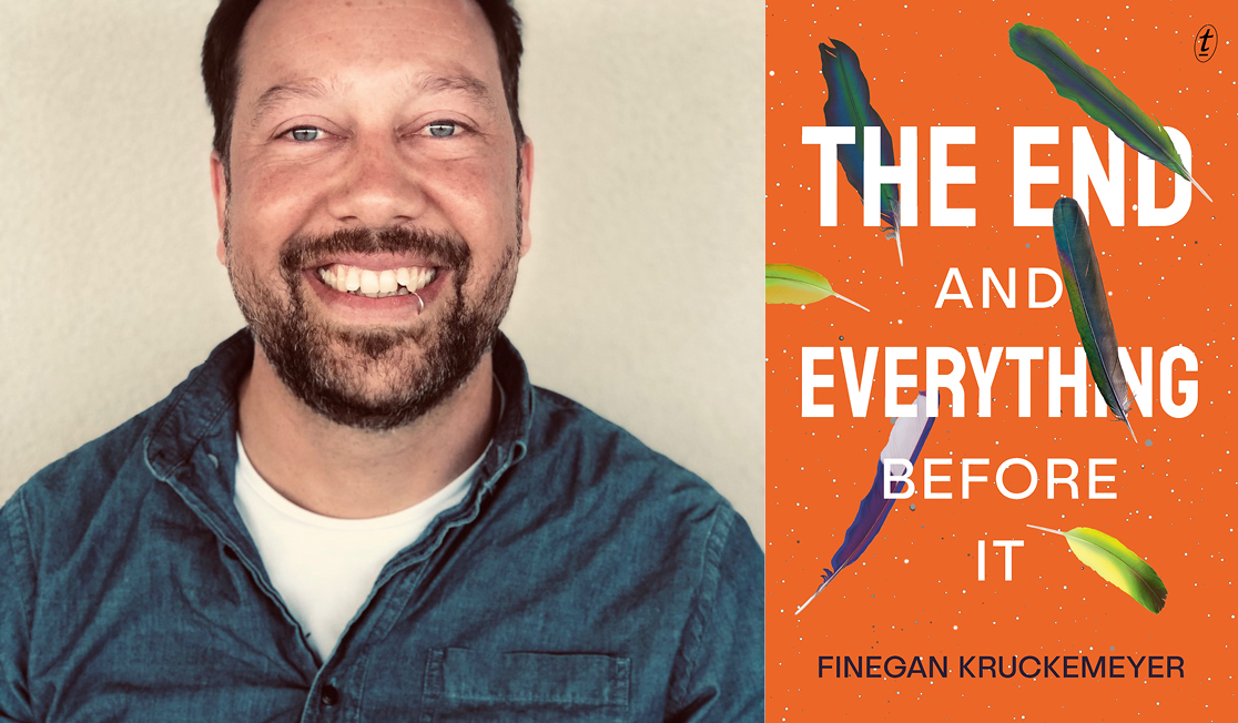 Finegan wears a blue shirt with a white t-shirt underneath. He has short dark hair and a short dark beard