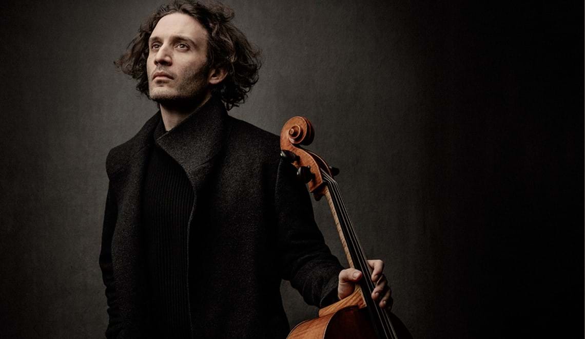 A musician stands in front of a dark background. He has medium length, dark, curly hair is look up into a spotlight and wears a high necked coat. He holds the neck of his cello in one hand.