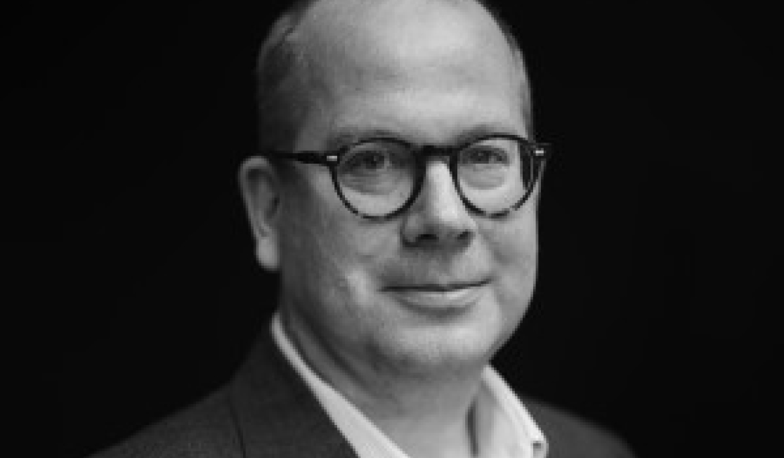 Tobias Buck is in front of a black background. Tobias wears glasses and a dark suit. He is smiling.