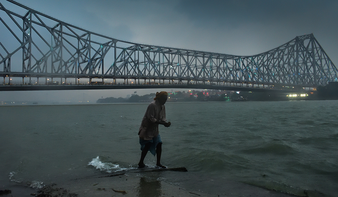 A photograph of a bridge over a body of water. A figure shrouded in darkness stands in the water, slightly bent over.