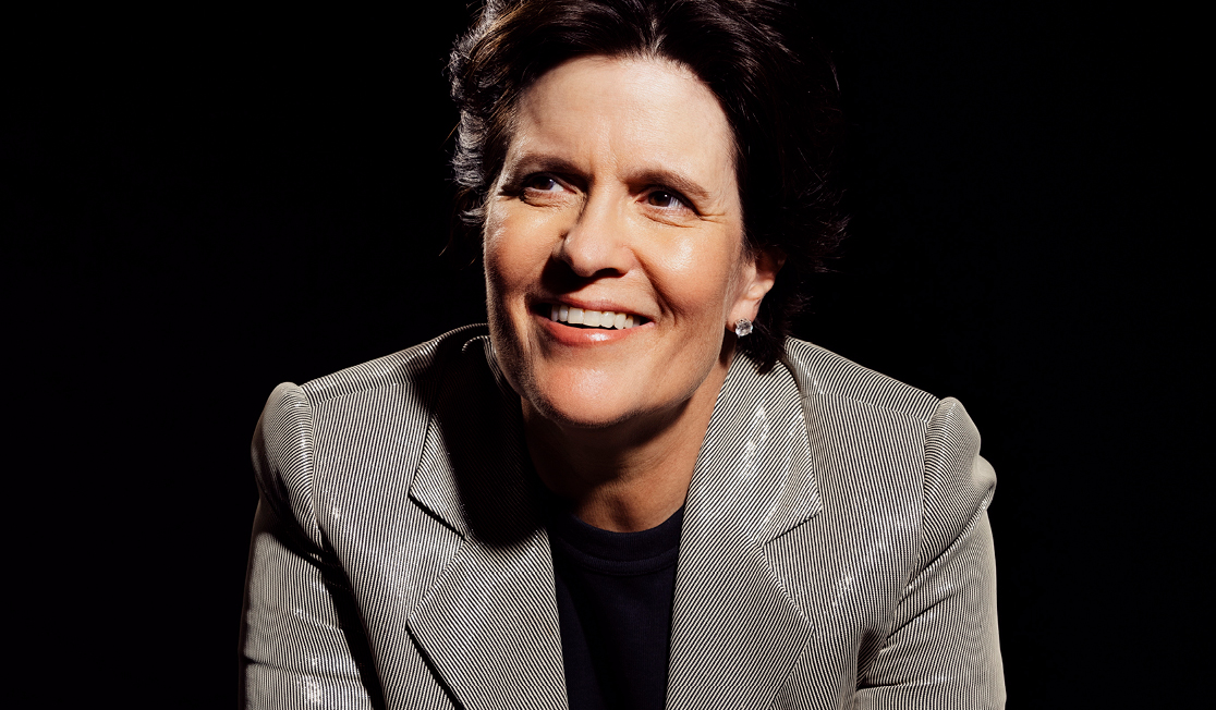 Kara Swisher wears a grey metallic suit and sits in front of a black background. Kara has short dark hair and is smiling.