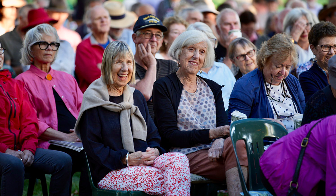 An audience sits on green chairs in an outdoor setting. There are two women smiling and watching what is happening on stage.
