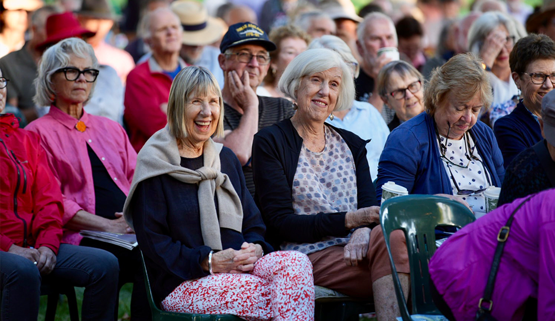 An audience sits on green chairs in an outdoor setting. There are two women smiling and watching what is happening on stage.