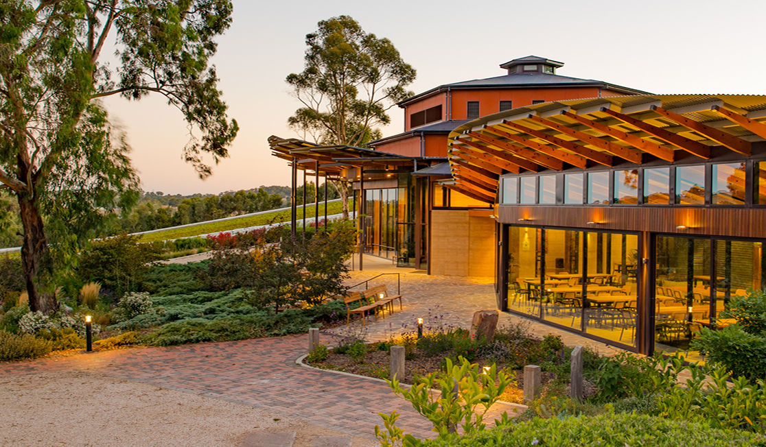 A modern looking building with lots of large windows surrounded by a garden, a path and gum trees. The sun is setting and the building is lit in golden light. There is also warm light coming from within.