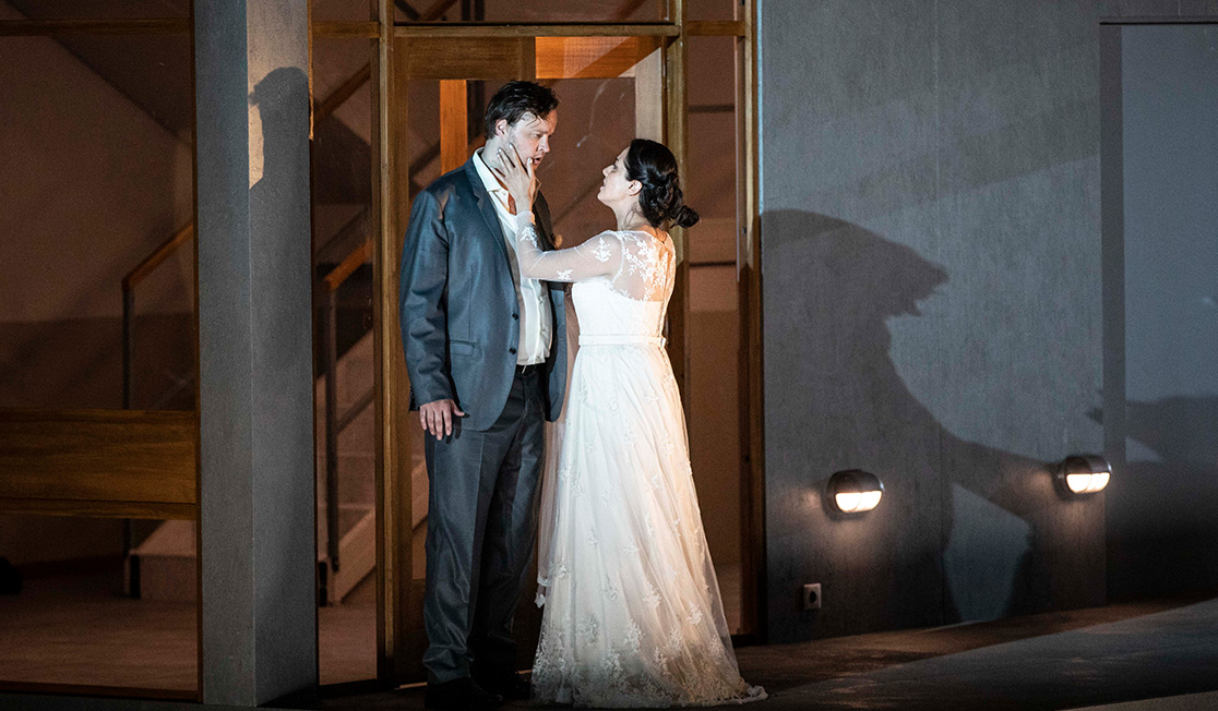 A two story building is set on stage. Inside we see people eating at tables in a restaurant, chefs in a kitchen and people in a bathroom. The lower floor is guests at a wedding reception dinner, there are heart balloons on the ceiling and guests are chatting.