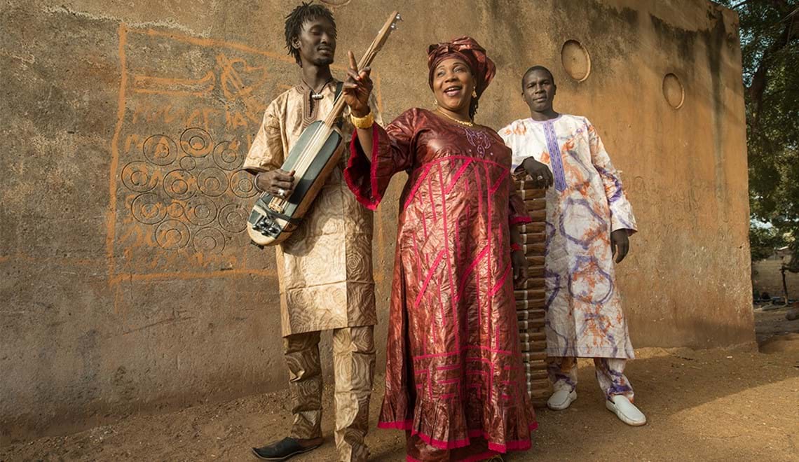 Three musicians stand in front of a cement wall, they are holding instruments and wearing clothes with intricate patterns. The woman in the middle is singing and has one arm raised.