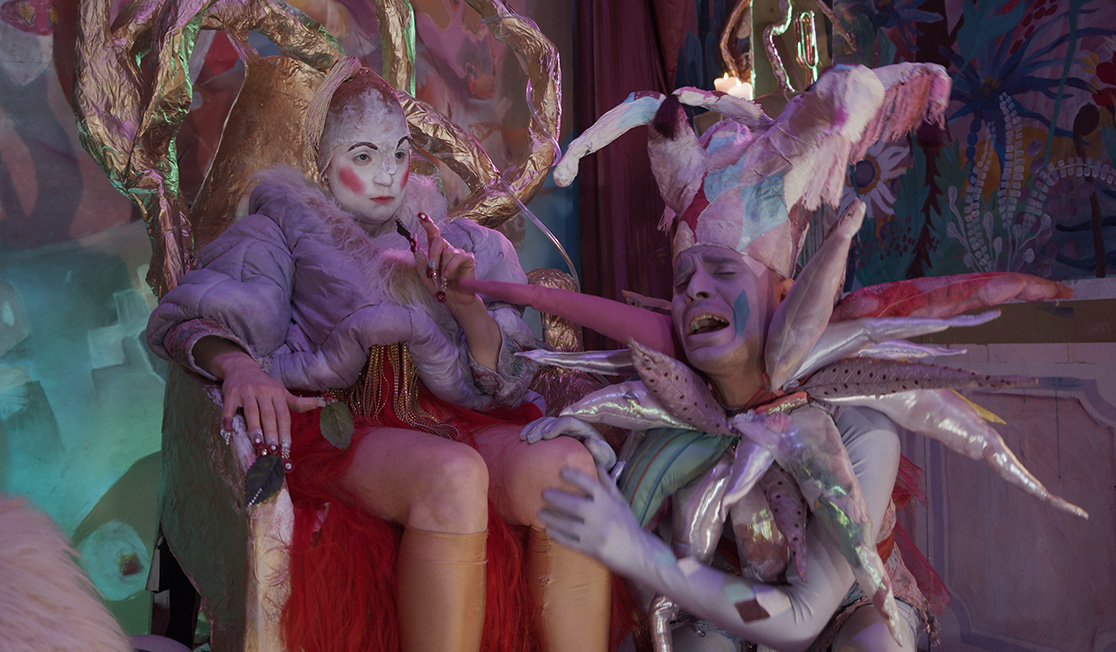 Two heavily costumed characters are in a dramatic pose. Both are wearing face paint and elaborate headgear. The first is sitting in a throne and the second appears to be crying while sitting at her feet.