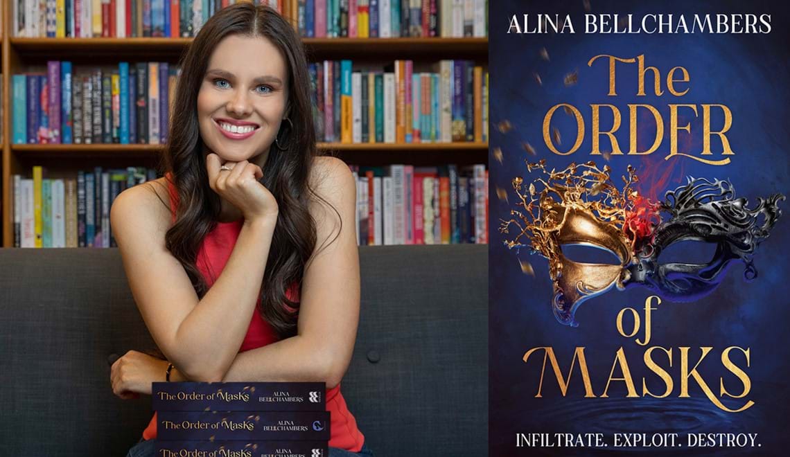 Alina Bellchambers wears a red dress and sits on a couch in front of a bookshelf. Alina has long brown hair and is smiling with one hand resting on their chin,