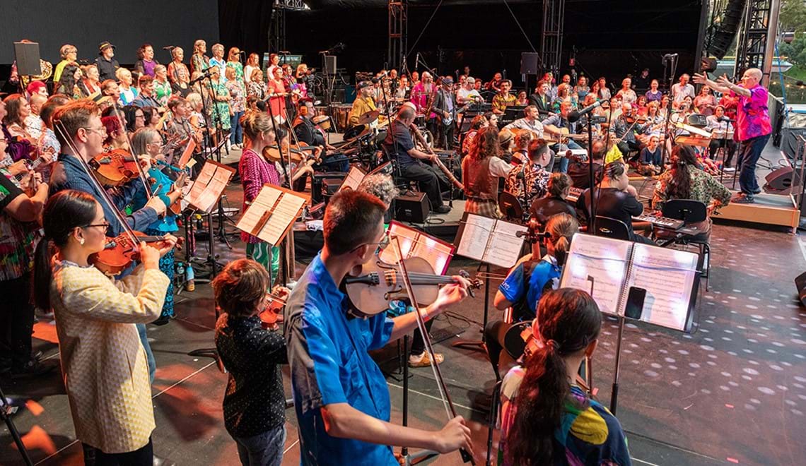 A large group of musicians of varying ages play on a stage while a man wearing a bright pink shirt conducts.