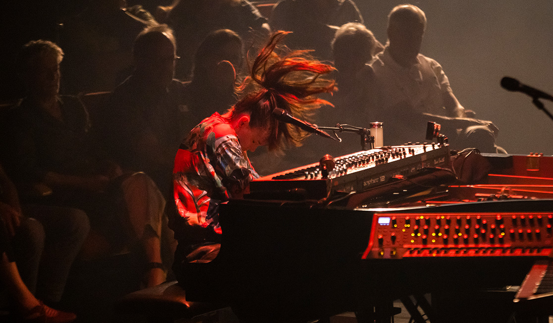 A woman is playing the piano, she's moving her head so that her hair falls forward. An audience is visible in the background.