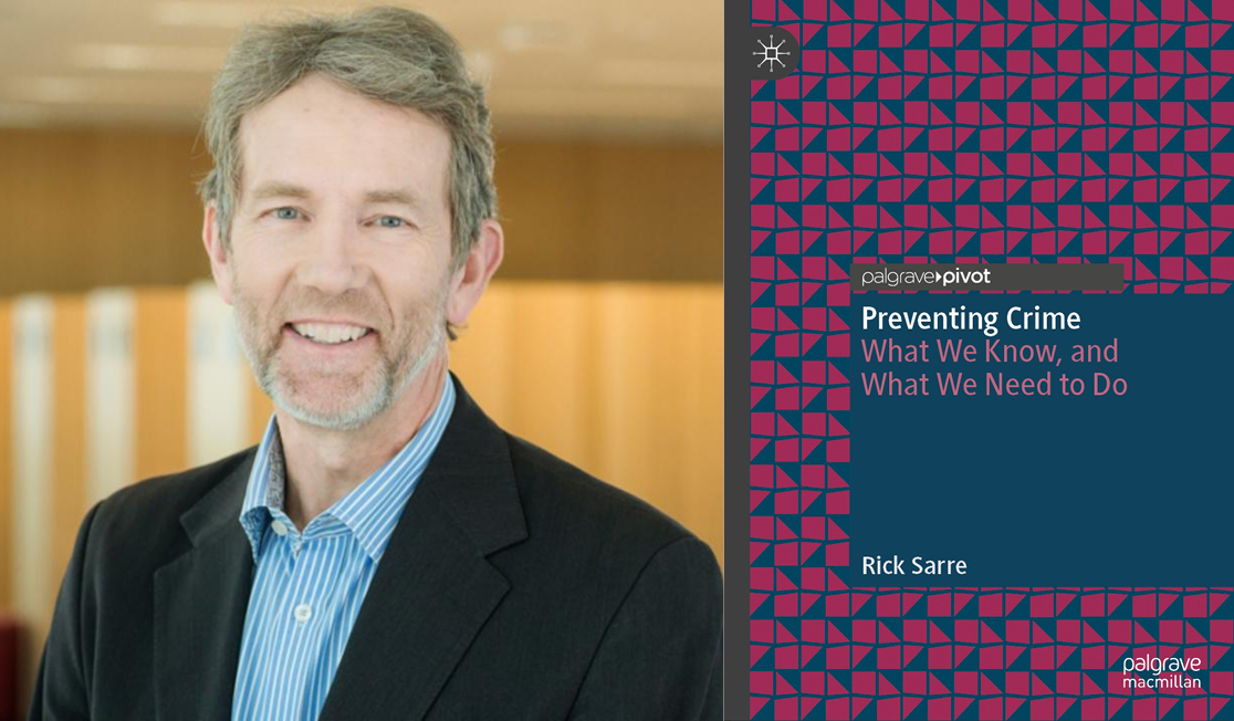 Rick Sarre wears a black suit jacket and a blue shirt. Rick has short grey hair and a beard and is smiling.