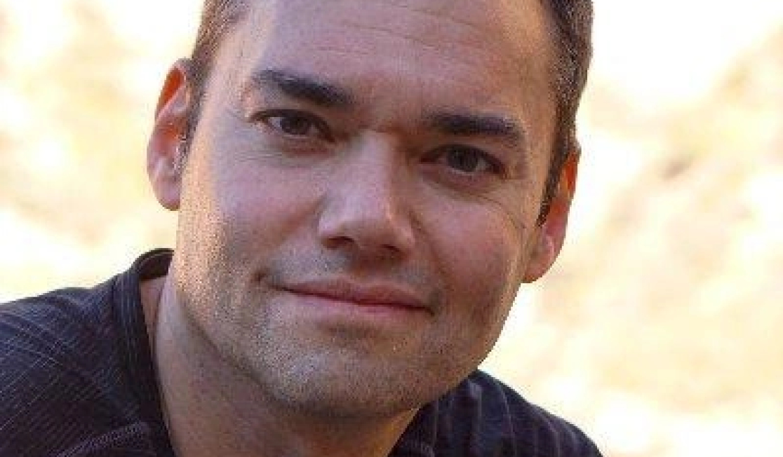 Peter Beinart has short dark hair and wears a black top. Peter is smiling at the camera and the background behind him is bright.