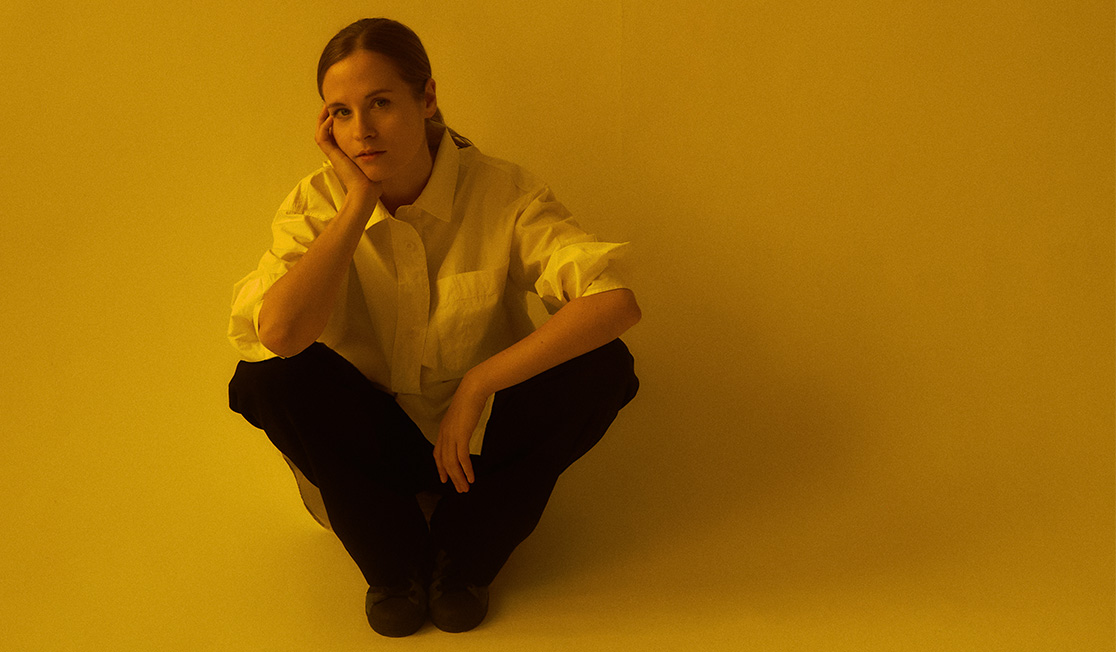 Hania sits on a white background wearing black pants and a white shirt. They have their head resting on one hand